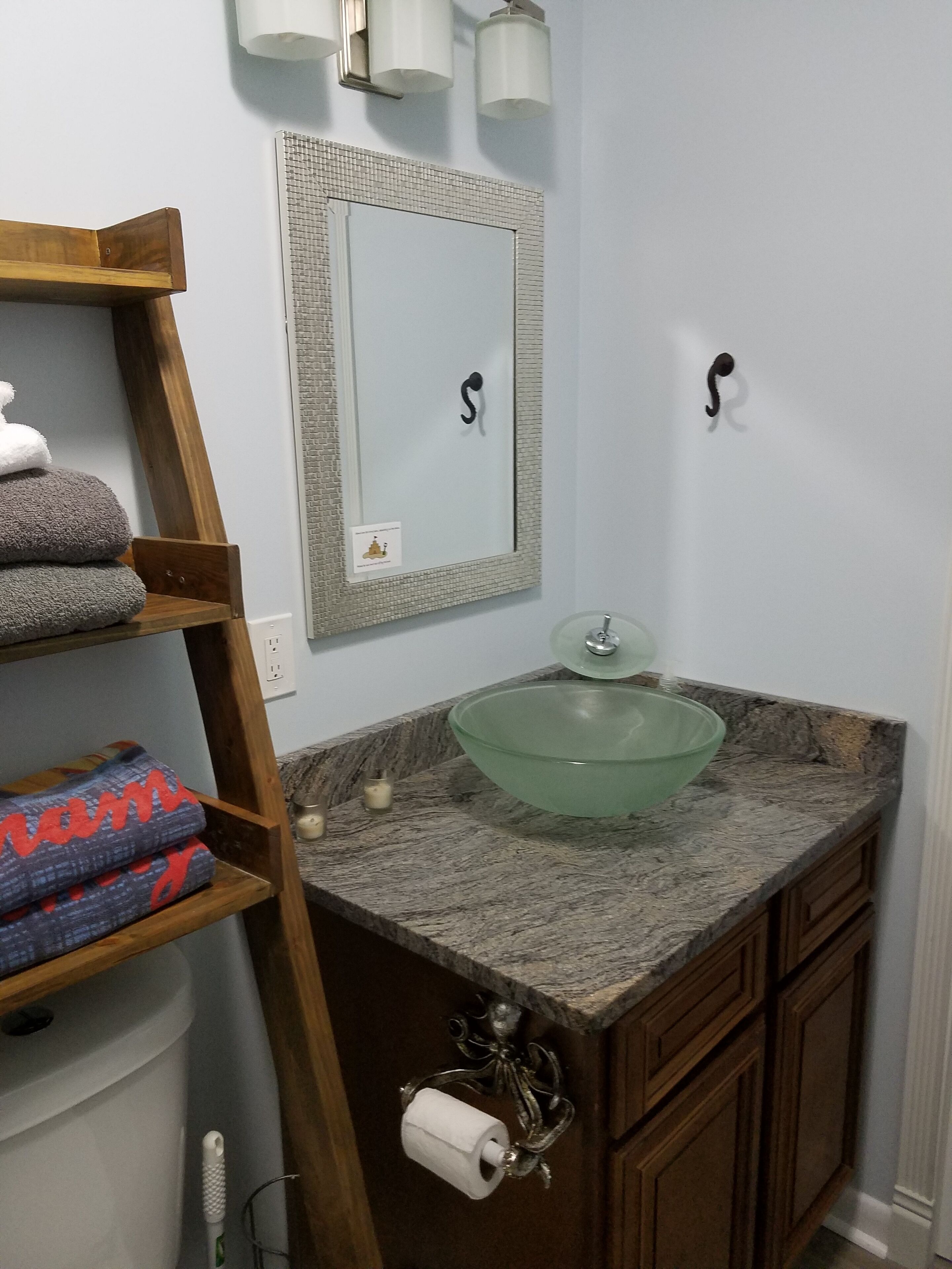 Une salle de bain avec une étagère en bois où sont posées des serviettes, un lavabo en verre et un miroir doté d’un cadre à motifs.