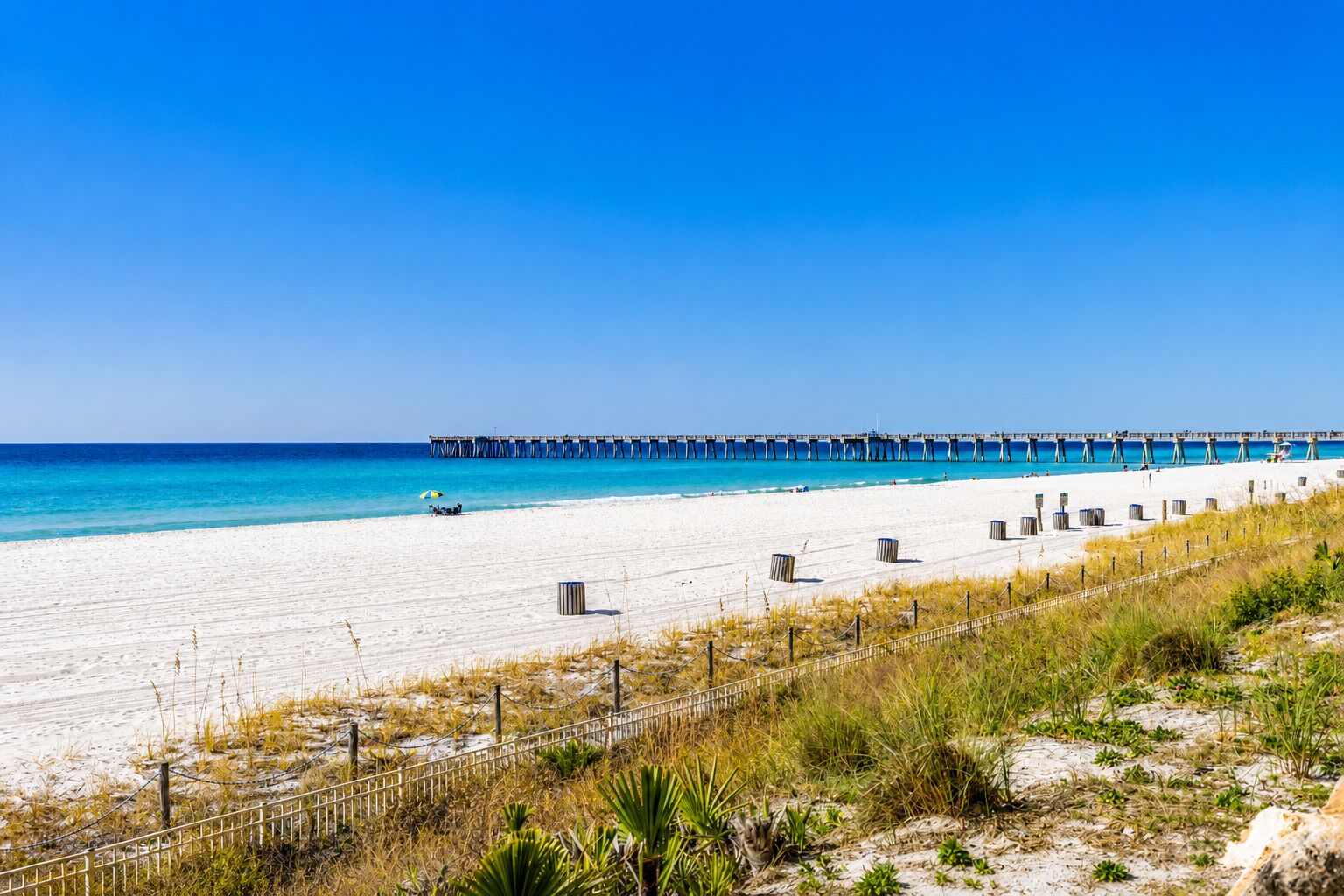 A beach with a pier extending into the ocean, sandy beach, and a fence in the foreground.