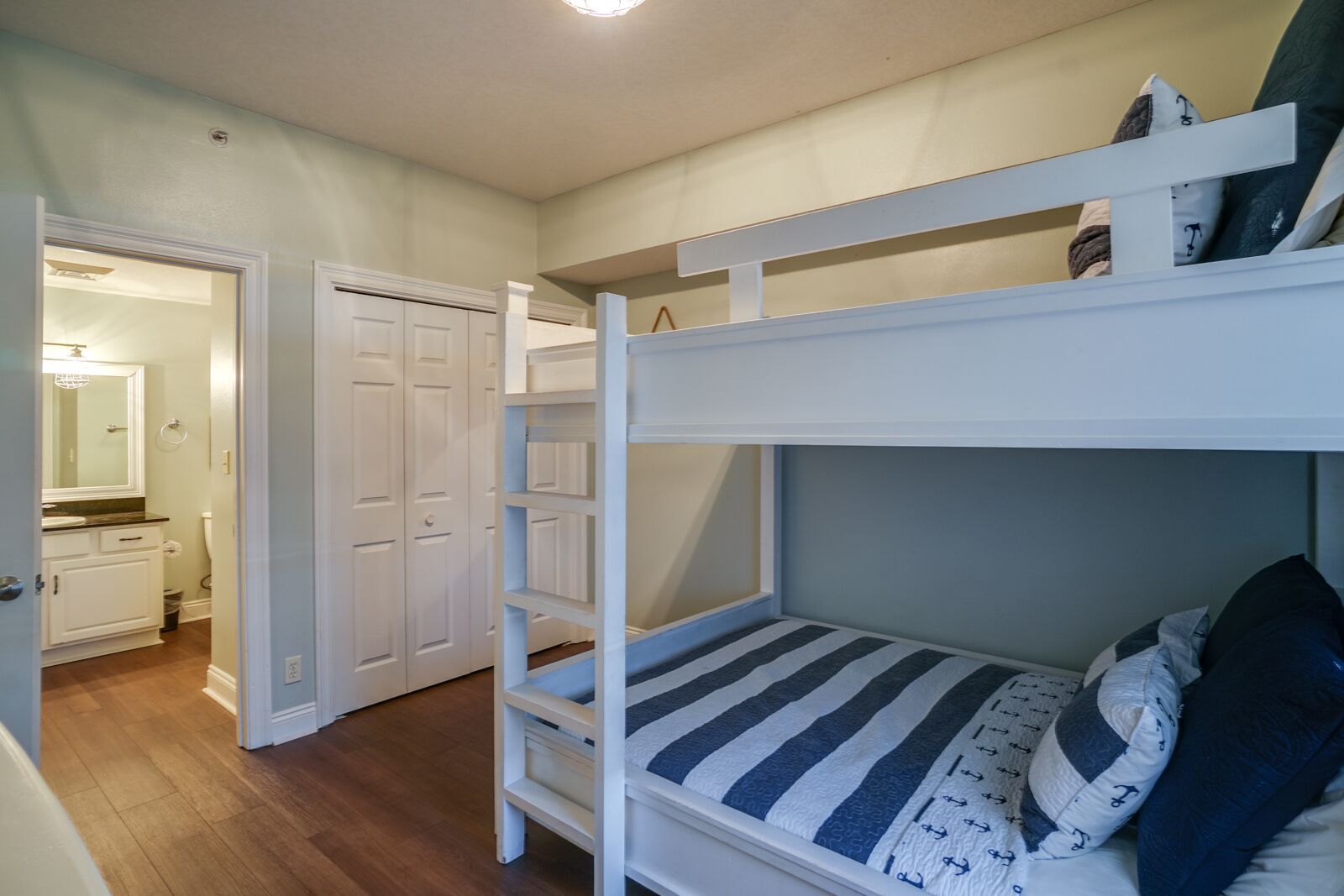 Una cama litera con colchón y almohadas a rayas, una puerta blanca y un baño con espejo y lavamanos.