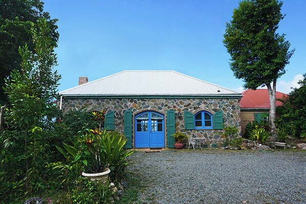 A stone building with a blue door and green shutters, surrounded by lush greenery.