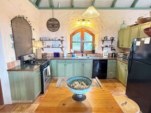 A kitchen with a stove, sink, and refrigerator. There is a window with a wooden frame and a hanging light fixture.