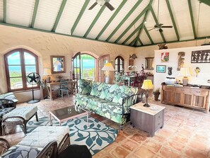 A spacious living area with a sofa, coffee table, and dining table. There are windows with views, a ceiling fan, and a wooden cabinet.