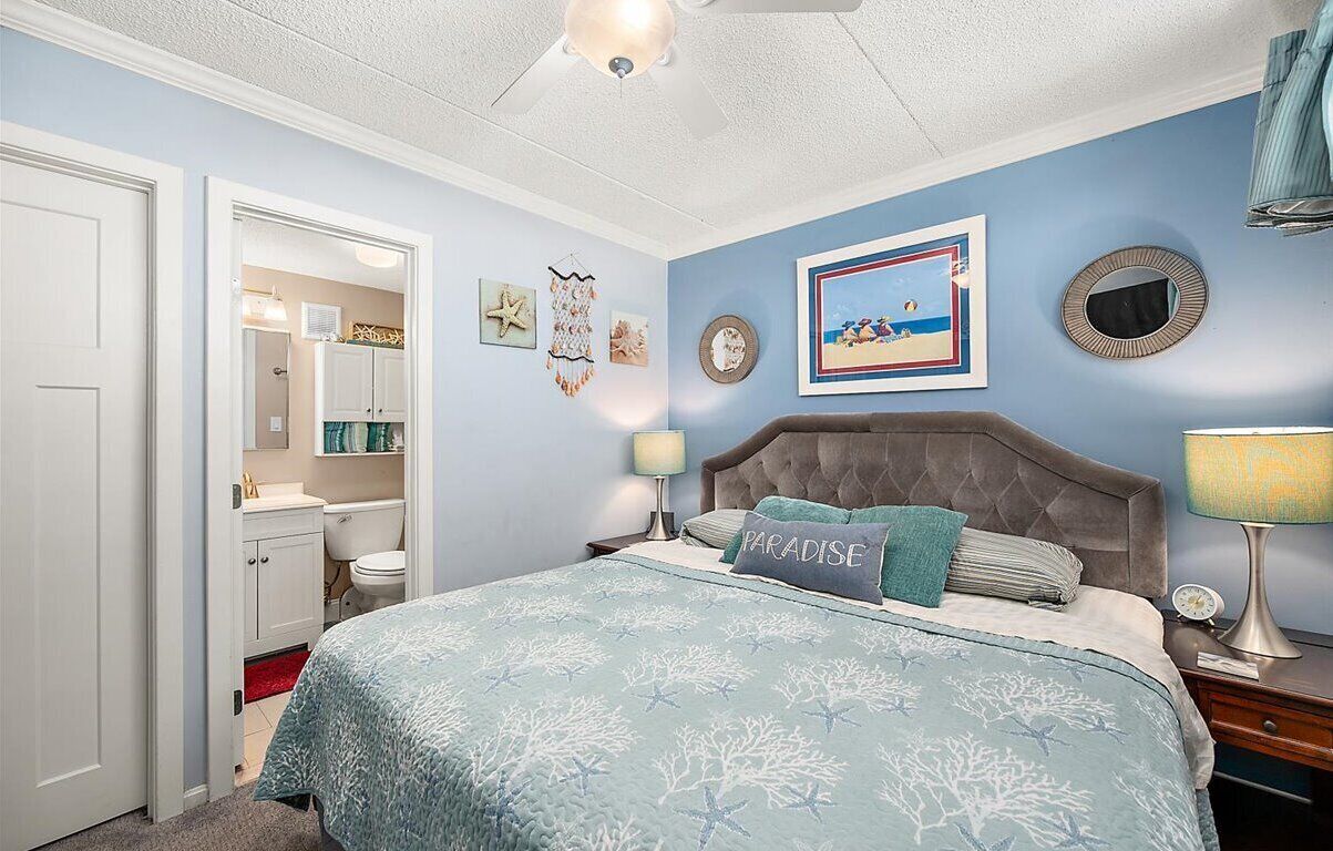 Ein Schlafzimmer mit einem Bett, einem Nachttisch, einer Wandlampe, einem Deckenventilator, einem gerahmten Bild und einem Spiegel.
