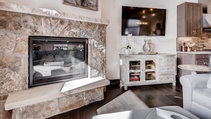 A modern living room with a stone fireplace, a flat-screen TV, a kitchen area with wooden cabinets, and a grey sofa. - Keystone 4BR Townhome at River Run Base - Prices lowered for open dates! (Keystone)