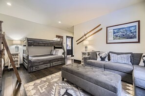 A modern living room with a bunk bed, grey sofa, and ottoman. There is a ski rack on the wall and a framed picture above the sofa.