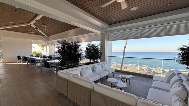 Un vaste espace intérieur doté d’un plafond en bois, d’un coin salon avec des canapés blancs et d’une table à manger, et offrant une vue sur la plage grâce à de grandes fenêtres.