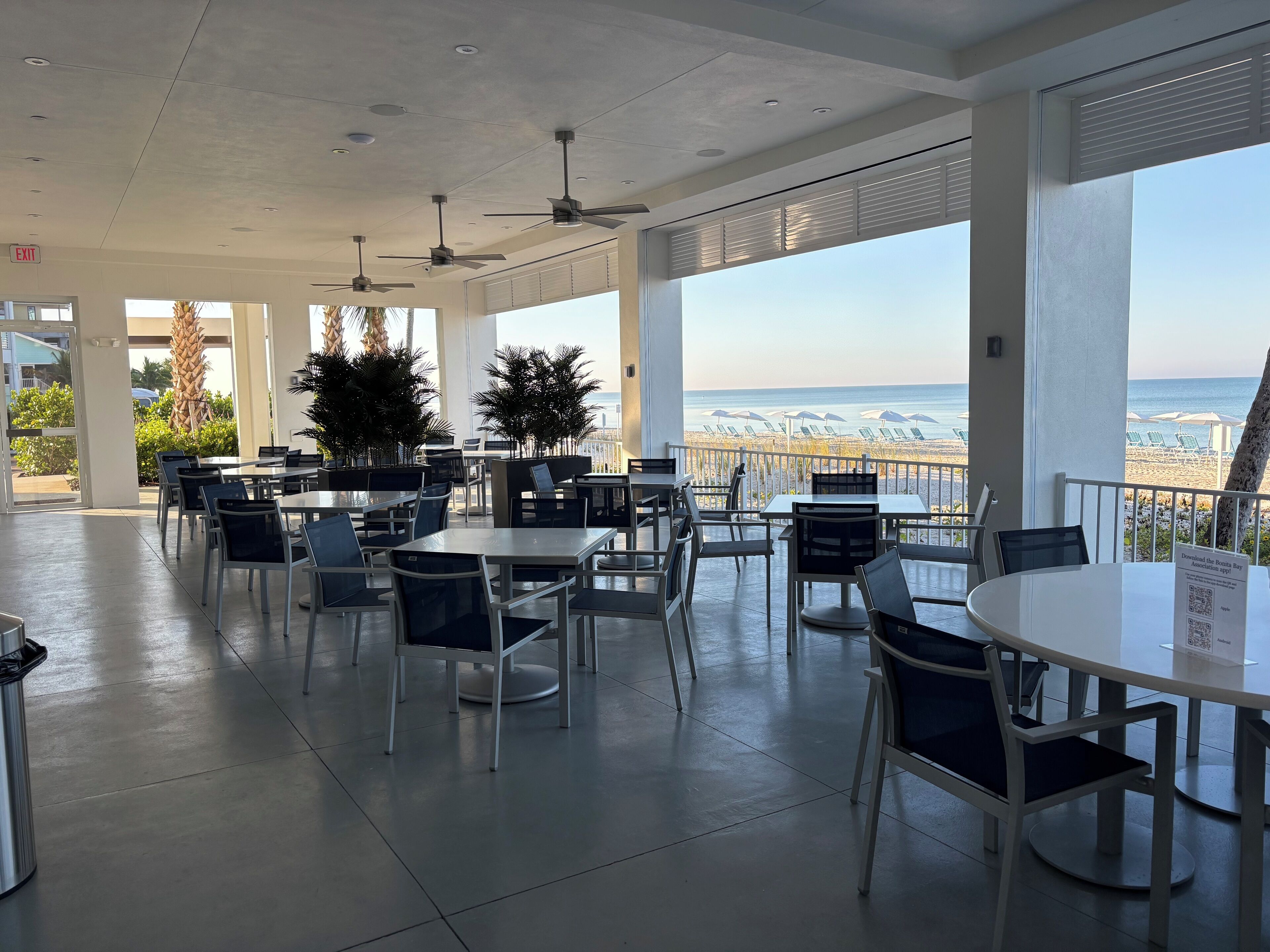 A spacious outdoor dining area with tables and chairs, large windows offering a view of the beach, ceiling fans, and a sign for a mobile app.
