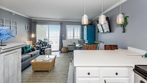 A modern living room with a sofa, coffee table, and dining area. There is a kitchen with white cabinets and a stove. The room has large windows and a balcony. - BEST DEAL ON THE KEY! Updated Gulf Front Condo! Full Ocean Views! (Pensacola)