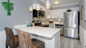 A modern kitchen with white cabinetry, a stainless steel refrigerator, and a central island with woven chairs. - BEST DEAL ON THE KEY! Updated Gulf Front Condo! Full Ocean Views! (Pensacola)