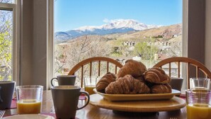 A breakfast setting with croissants, a coffee cup, and juice glasses, overlooking a mountainous landscape. - Pikes Peak Views! Fire Pit, Pool Table & Arcade ~ 4 King Beds, Dogs OK! (Colorado Springs)