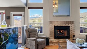 A living room with a fireplace, a comfortable armchair, a large window with a mountain view, and a painting on the wall. - Pikes Peak Views! Fire Pit, Pool Table & Arcade ~ 4 King Beds, Dogs OK! (Colorado Springs)
