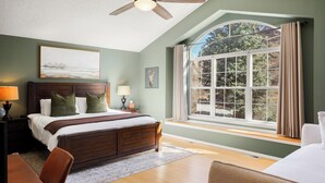 Ein Schlafzimmer mit einem großen Bett, einem Deckenventilator, einem Fenster mit Vorhängen und Blick auf Bäume.