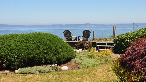 Une vue d’un jardin avec deux chaises Adirondack donnant sur un plan d’eau, une terrasse en bois et une montagne à l’horizon.