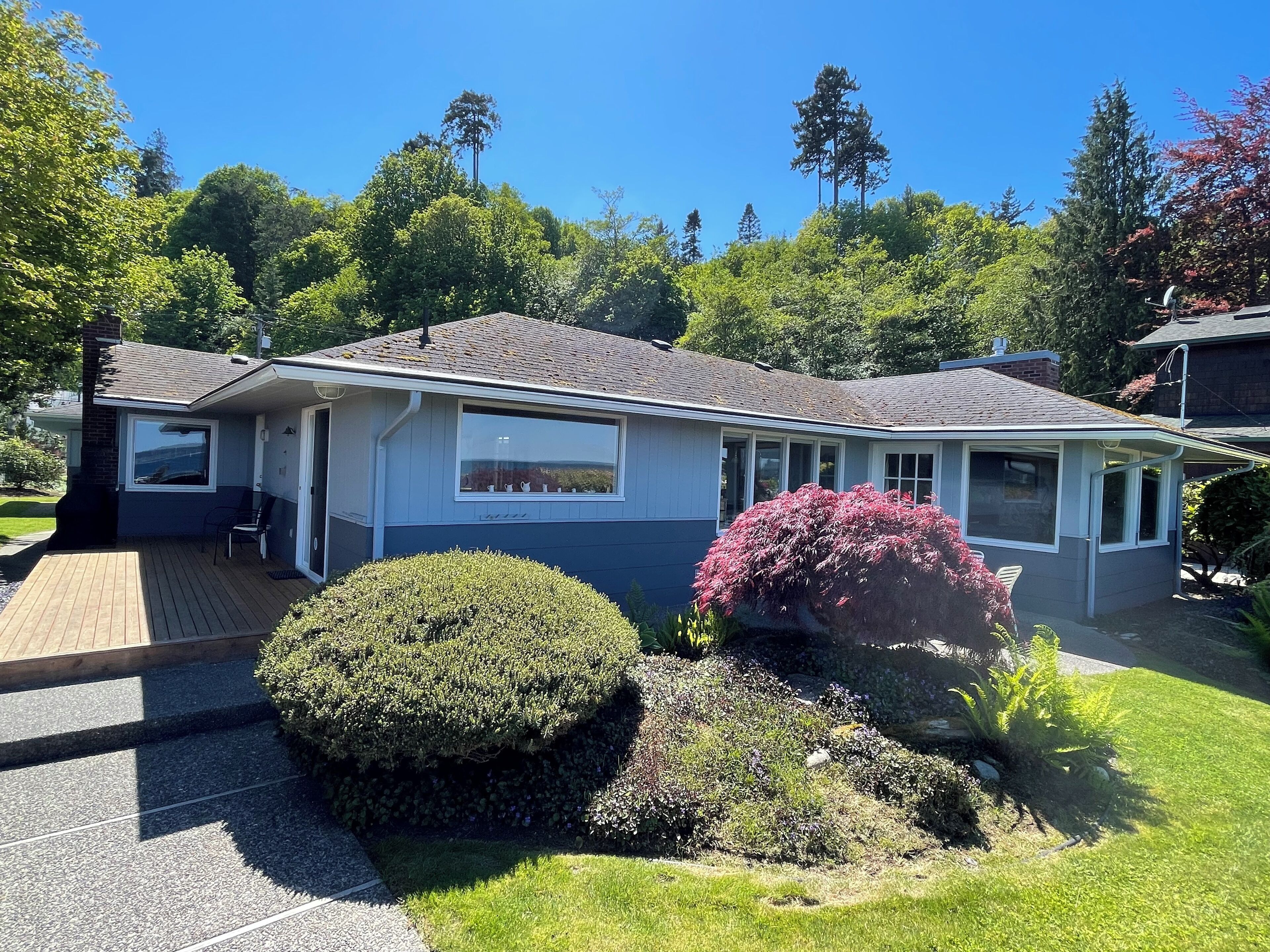 A single-story house with a covered porch, a small garden with a bush and a flowering plant, and a clear blue sky.