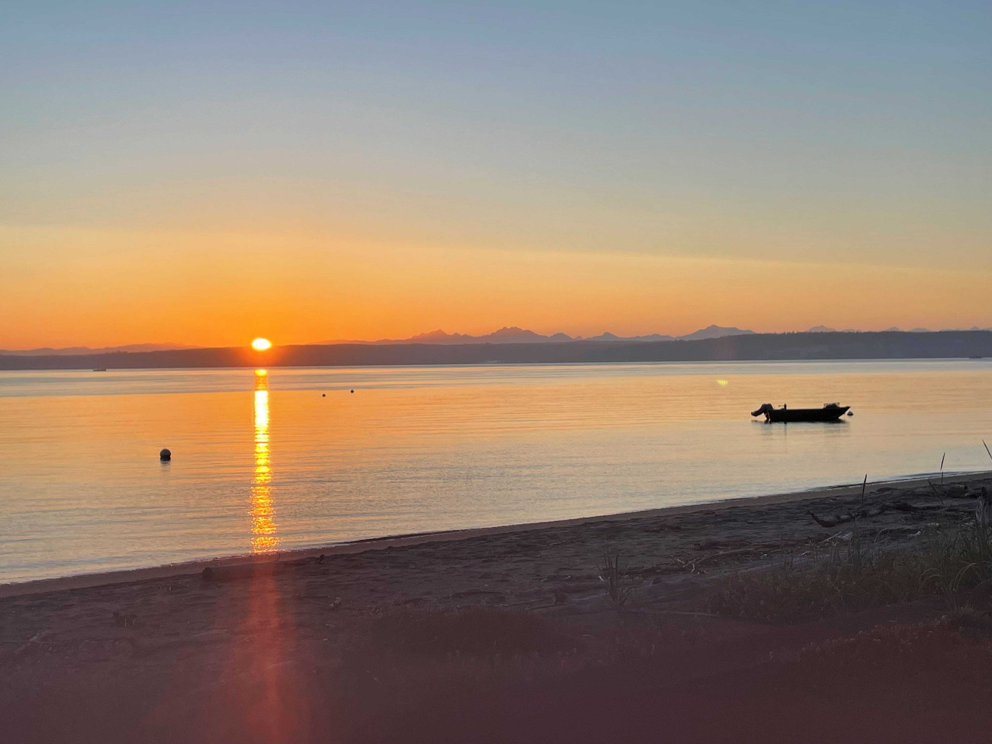 Un coucher de soleil sur une étendue d’eau calme, avec un bateau et des montagnes au loin.