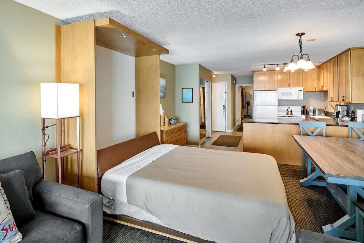 A hotel room with a bed, a desk, a chair, a lamp, and a kitchenette.