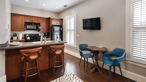 Una cocina moderna con armarios de madera, un refrigerador de acero inoxidable y un microondas integrado. Hay una barra con taburetes y una pequeña mesa con sillas. Un televisor de pantalla plana está montado en la pared.