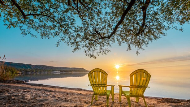 Two chairs facing a sunset over a lake with a tree overhead.