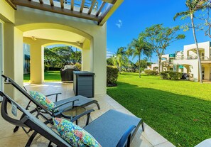 A covered outdoor seating area with lounge chairs and a trash bin, a well-maintained lawn, and a building with multiple balconies.
