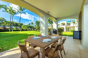 An outdoor patio with a table set for dining, surrounded by wicker chairs, and overlooking a well-maintained lawn with palm trees.