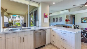 A modern kitchen with a dishwasher, a large window, and a dining area with chairs. - Rare No-Stairs Ocean View Condo ~ Palms at Wailea (Wailea)