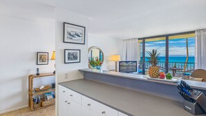 A modern kitchen with a view of the ocean, a dining area with a table and chairs, and a counter with a fruit display. - Oceanfront Condo at Sugar Beach Resort- Stunning Sunsets and Beach Front (Kihei)