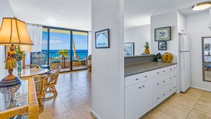 A modern kitchen with white cabinets, a dining area with wicker chairs, and a large window overlooking the ocean. - Oceanfront Condo at Sugar Beach Resort- Stunning Sunsets and Beach Front (Kihei)