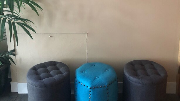 Eine Wand mit einem Schild, auf dem steht "YOU ARE NOW ENTERING A STRESS FREE ZONE", und zwei Fischdekorationen. Vor der Wand befinden sich drei Hocker, ein blauer und zwei graue.