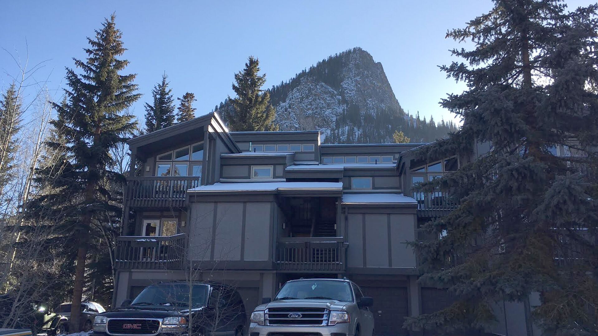 Un edificio de varios pisos con un balcón, rodeado de altos árboles y una montaña al fondo.