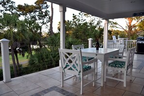 Un patio couvert avec une table et des chaises blanches, un barbecue et une vue sur un jardin aménagé.