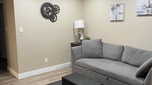 Un salon avec un canapé gris, un pouf noir, une horloge murale, une lampe et deux tableaux.