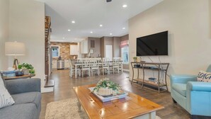 A modern living room with a wooden coffee table, a grey sofa, and a blue armchair. A flat-screen TV is mounted on the wall, and a dining area with white chairs is visible in the background. - 22 Grover's Ocean, spacious 5 bedroom home with central AC, 1 block to beach! (Ocean Grove)