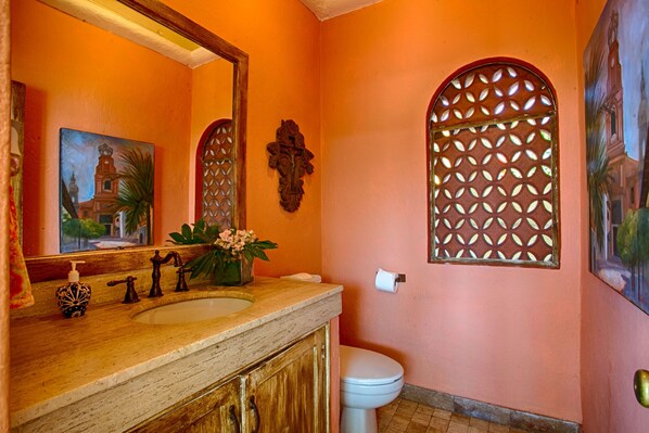 A bathroom with a sink, mirror, toilet, decorative window, and a painting on the wall.