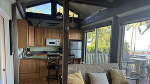 A cozy living area with a sofa and a wooden coffee table, a kitchen with wooden cabinets and a microwave, and a large window offering a view of palm trees and the ocean. - Private Oceanfront Makena Retreat for 2, Maui County Permitted Short-Term Rental (Kihei)