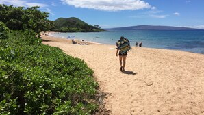 A sandy beach with a clear sky, a hilly landscape in the distance, and a person carrying a backpack walking along the shore. - Private Oceanfront Makena Retreat for 2, Maui County Permitted Short-Term Rental (Kihei)
