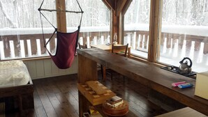 A cozy cabin interior with a wooden dining table, chairs, a hanging chair, a small kitchen area with a kettle, and a view of a snowy forest through large windows. - Mökki Hovland Hut - A Unique Glass Wall Timber Frame Nestled in a Maple Forest (East Cook)