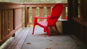 A wooden deck with a red chair facing a forested area. - Mökki Hovland Hut - A Unique Glass Wall Timber Frame Nestled in a Maple Forest (East Cook)