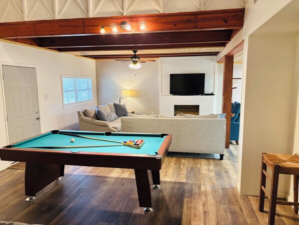 A pool table in a room with a sofa, a television above a fireplace, and wooden beams on the ceiling.