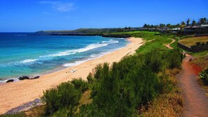 Paesaggio costiero con spiaggia sabbiosa, cielo azzurro limpido e un sentiero che conduce a un edificio.
