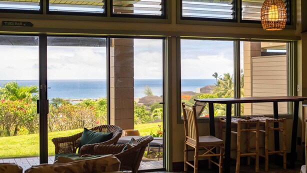 Una stanza con ampie porte scorrevoli in vetro che si aprono su un balcone vista oceano. Sul balcone, una sedia in vimini e un tavolo con sedie.