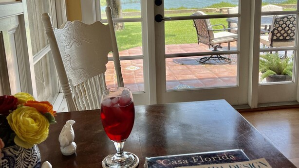 A table with a glass of red drink, a magazine, and decorative items. A white chair is next to the table, and a view of a body of water and greenery is visible through a glass door. - An irresistable oceanfront vacation home in the heart of historic St Augustine! (St Augustine)