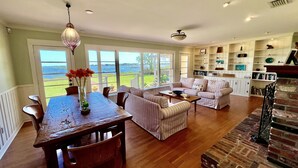 A spacious living room with a large window, a dining area with a wooden table, and a brick fireplace. - An irresistable oceanfront vacation home in the heart of historic St Augustine! (St Augustine)