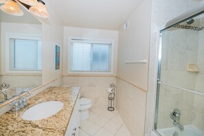 Een badkamer met een wastafel met granieten blad, toilet, douche met glazen deur en twee ramen met zonwering.