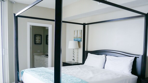 A four-poster bed with a white and teal bedspread, a nightstand with a lamp, and a door leading to another room. - Beach Colony West 3C/Gulf Front 3BR/3BA w/Beach Chair Service & WINTER RATES (Navarre Beach)