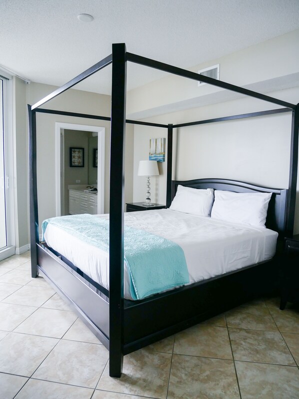 A four-poster bed with a white and teal bedspread, a nightstand with a lamp, and a door leading to another room. - Beach Colony West 3C/Gulf Front 3BR/3BA w/Beach Chair Service & WINTER RATES (Navarre Beach)
