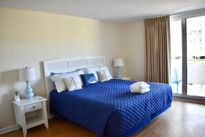 Quarto com colcha azul acolchoada, cabeceira branca e mesa de cabeceira branca com abajur.