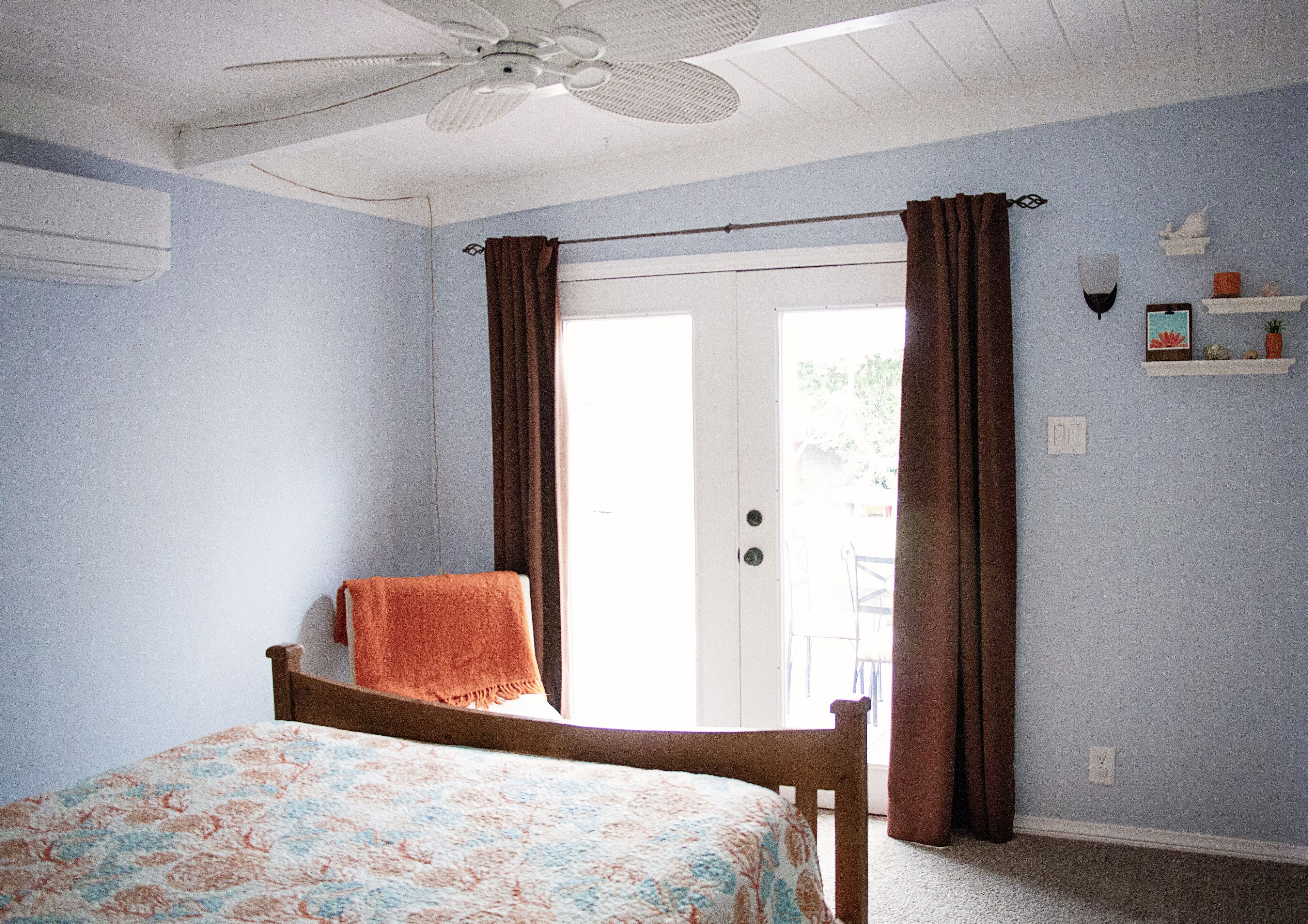 Une chambre à coucher avec un lit, un ventilateur de plafond, une porte avec des rideaux et une petite étagère ornée d’objets décoratifs.
