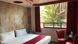 Quarto de hotel com uma cama grande, uma pequena mesa com cadeiras, uma janela com vista para palmeiras e uma parede de pedra.