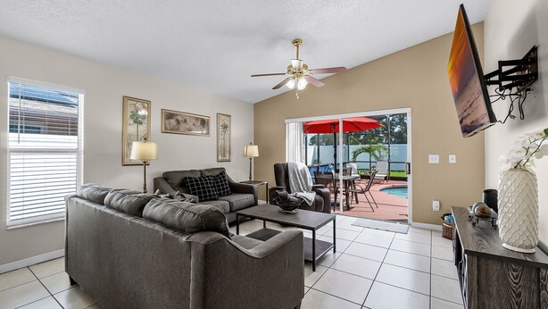 A living room with a gray sofa, a coffee table, and a ceiling fan. There is a sliding glass door leading to an outdoor patio with a pool. - BUENAVENTURA LAKES (263HS) - 3BR 2BA home with pool in a private setting (Kissimmee)