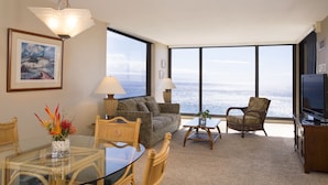 A spacious living room with a large window overlooking the ocean, a comfortable sofa, a dining table with chairs, and a television on a wooden stand.